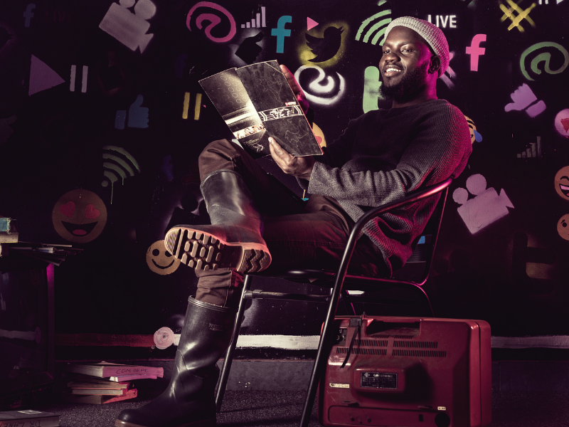 A man of African origin sitting on a chair.  He is wearing a beanie and holding a booklet.  Behind him are lots of colour social media icons.